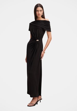 Femme aux longs cheveux lisses portant une robe noire épaules dénudées avec un détail froncé à la taille et des sandales à talons noires, se tenant sur un fond uni.