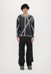 Grauer und schwarzer Pullover mit diagonalen hellgrauen Streifen, Rundhalsausschnitt und engen Bündchen, kombiniert mit lockeren schwarzen Cargo-Hosen und grünen Schuhen.