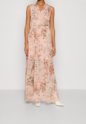 Ärmelloses pinkes Maxikleid mit Blumenmuster, V-Ausschnitt und dünnem geflochtenem Gürtel mit Quasten, getragen mit weißen spitzen Schuhen.