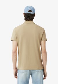 Camisa polo beige con una textura suave y mangas cortas, combinada con una gorra de mezclilla azul clara. El modelo se muestra de espaldas.