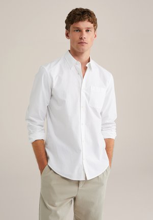 Camisa blanca de botones con un bolsillo de parche, tejido, cuello puntiagudo y mangas arremangadas, combinada con pantalones beige claros.