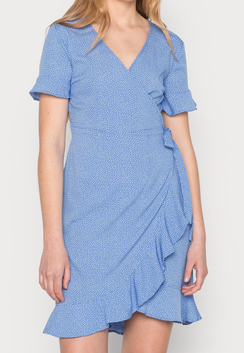 Robe portefeuille bleue à pois blancs, manches courtes à volants, décolleté en V et ourlet asymétrique à volants sur une personne debout.