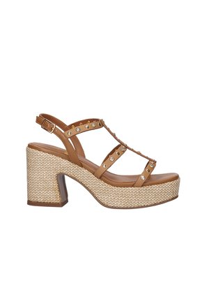 Sandalia de plataforma de cuero marrón con tacón grueso tejido, tira en T adornada con tachuelas doradas y plateadas, y hebilla ajustable en el tobillo.