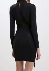 Kvinna klädd i en figursydd svart glittrig långärmad miniklänning med hög hals och en liten knappstängning i ryggen.