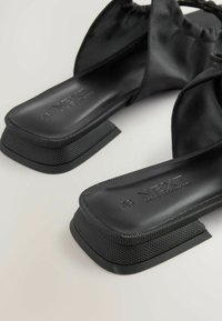 Sandales noires avec une semelle en caoutchouc texturé, arborant un design élégant et des sangles en tissu doux. La semelle intérieure est embossée du logo "NEXT".