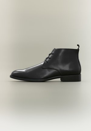 Bottine en cuir noir avec lacets, bout rond, petit talon et languette à l'arrière sur un fond clair uni.