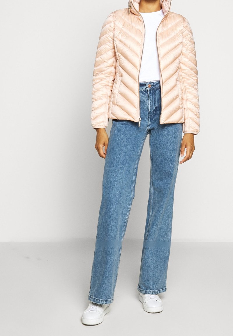 Personne portant une veste matelassée rose clair, un t-shirt blanc, un jean bleu taille haute et des baskets blanches, debout devant un fond uni.