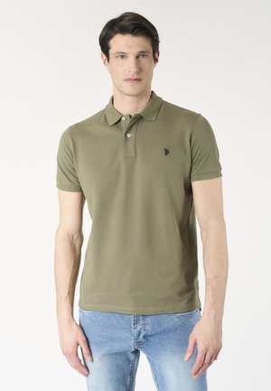 Uomo che indossa una polo verde oliva a maniche corte con piccolo logo sul petto e jeans azzurri, in piedi davanti a sfondo bianco.