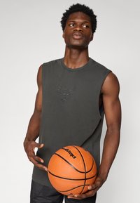 Ermeløs mørkegrå tank topp med en brodert okselogo på brystet. Modellen holder en oransje basketball.