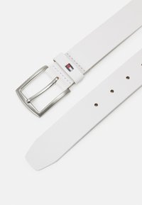 White leather belt with a rectangular silver buckle. Features a logo patch in red and blue. Smooth texture and pointed end design.