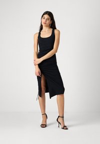 Svart, ermeløs midikjole med samlet detaljer langs siden og en høy splitt. Glatt stoff, tettsittende fasong, minimalistisk design. Par med svarte høyhælede sko.
