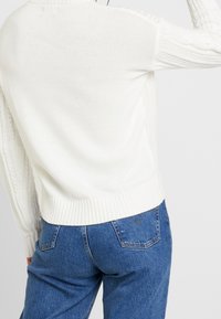Person wearing white knitted sweater and blue denim jeans, shown from the back against a plain light background.