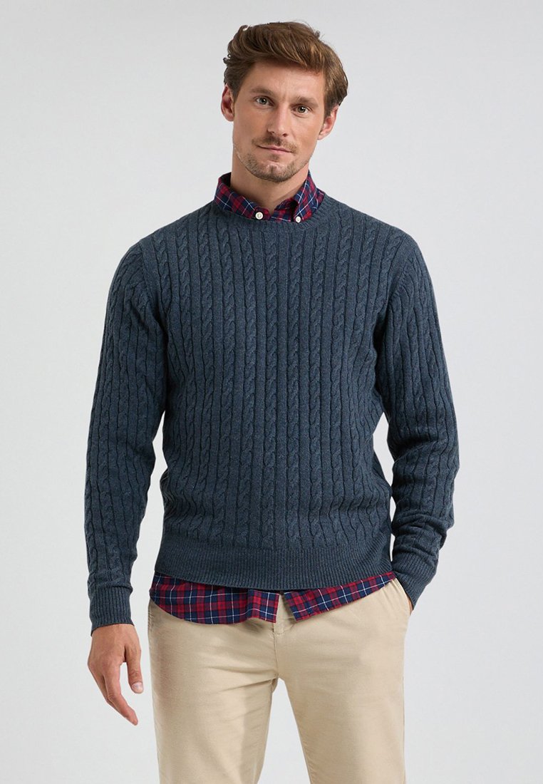 Donkerblauwe kabelgebreide trui met een ronde hals en geribde manchetten, gedragen over een rood-blauw geruit overhemd. Lichtbeige broek.