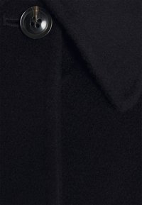 Gros plan d'un manteau en laine noire montrant un tissu texturé, un grand bouton foncé à quatre trous et un col pointu.