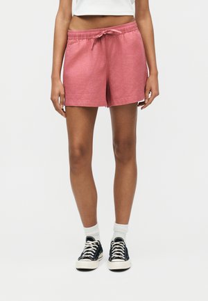 Személy rózsaszín zsinóros rövidnadrágot, fehér zoknit és fekete alacsony szárú tornacipőt visel, és egyszínű fehér háttéren áll.