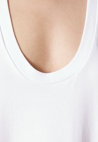Bílý bavlněný top na ramínka s širokým, žebrovaným kulatým výstřihem. Hladká textura a čisté okraje, které ukazují minimalistický design.