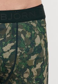 Boxers en tissu camouflé dans des nuances de vert et de beige, avec une ceinture élastique vert foncé portant le nom de la marque en relief.
