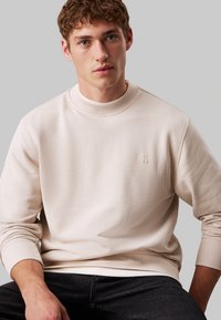 Felpa beige chiaro con colletto e polsini a costine texturizzati, caratterizzata da un piccolo logo in rilievo sul petto, abbinata a jeans scuri.