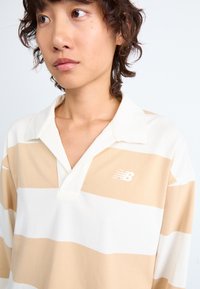 Beige och vit randig polo skjorta med V-hals och en vit logotyp. Tyget verkar vara lätt med en mjuk textur.