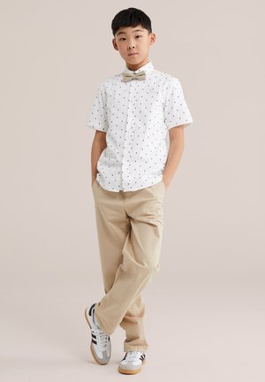 Chlapec stojaci s rukami v kapsách, oblečený v bielej košeli s krátkym rukávom a malými čiernymi vzormi, béžové nohavice, motýlik a tenisky.