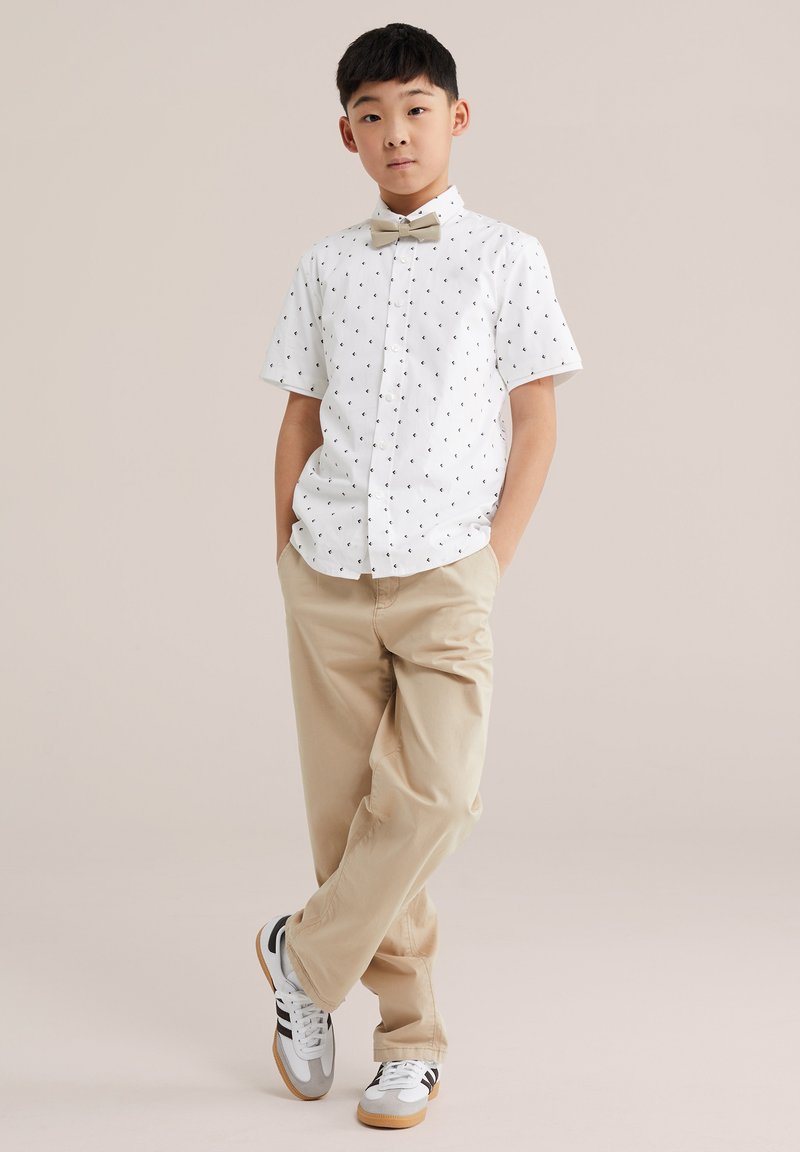 Poika seisoo käsineen taskuissa valkoinen lyhythihainen paita, jossa on pieniä mustia kuvioita, beigehousut, rusetti ja tennarit yllään.