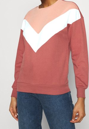 Personne portant un sweat-shirt à manches longues à blocs de couleurs rose poudré, blanc et pêche, associé à un jean en denim bleu.