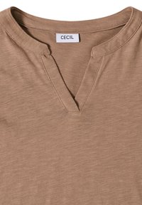 Lysbrun blød stof-top med rund halsudskæring, der har en lille V-formet klip og et hvidt mærke med teksten "CECIL" ved kraven.