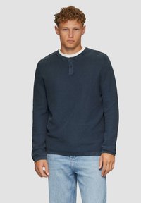 Granatowa koszula typu henley z długim rękawem, wykonana z teksturowanej tkaniny, z dwoma guzikami przy dekolcie i kontrastowym białym okrągłym dekoltem. Zestawiona z jasnoniebieskimi dżinsami.