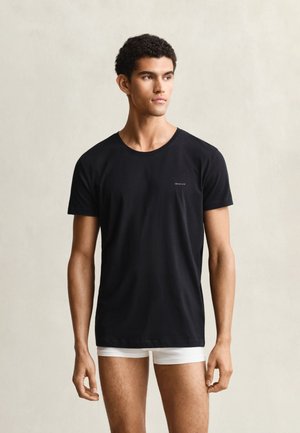 Jeune homme portant un t-shirt noir à manches courtes et un boxer blanc, debout devant un fond clair uni.