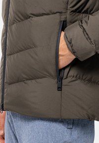Giacca a vento verde scuro con cuciture orizzontali, tasca laterale con zip e una finitura liscia e opaca. Indossata su pantaloni blu con texture.