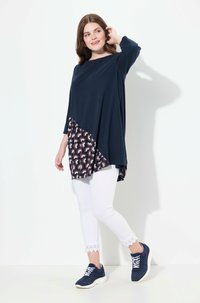 Mörkblå topp med asymmetrisk kant och mönstrad nederdel, i kombination med vita leggings med spetsdetalj och mörkblå sneakers.