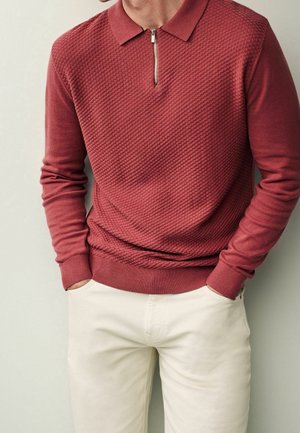 Homme portant un polo à manches longues rouge rouille texturé avec col zippé et pantalon blanc cassé, mains dans les poches, sur fond clair uni.