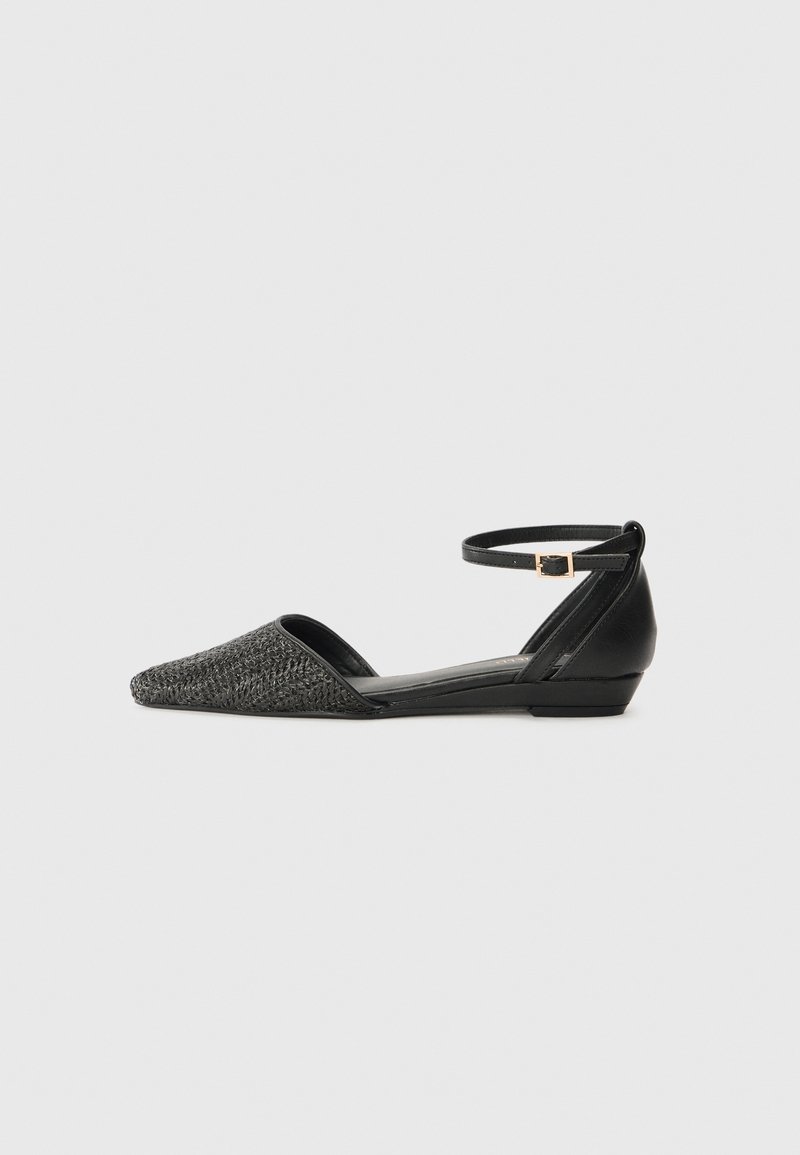 Chaussure plate noire avec bout pointu tissé, côtés ouverts, talon fermé et bride à la cheville avec petite boucle dorée sur fond gris.
