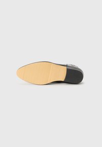 Suela de zapato de cuero negro con una parte inferior de goma marrón texturizada y un tacón negro liso, con un logotipo grabado en el lateral.