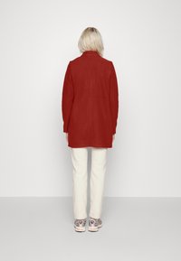 Chaqueta roja larga con corte recto, un cuello y una textura suave, combinada con pantalones color crema y zapatillas grises. Vista trasera.