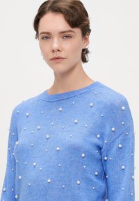 Lyseblå strikkegenser med rund hals og spredte hvite perlepynt mot et teksturert stoff.