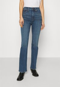 Kvinna klädd i högmidjade blå raka jeans i kombination med svarta ankelstövlar, stående mot en ljus enfärgad bakgrund.