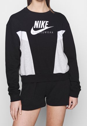 Woman wearing black, white, and gray Nike sportswear sweatshirt with logo, paired with black shorts, standing against plain background.