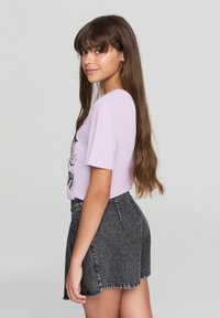 T-shirt a maniche corte di colore viola chiaro con stampa grafica, abbinato a pantaloni corti in denim neri a vita alta. Orlo curvo e dettagli di sfilacciatura discreti.
