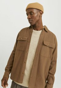 Brown button-up shirt with two chest pockets, layered over a light beige sweater. The model wears a tan ribbed beanie.