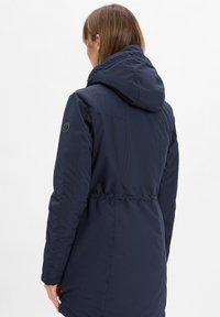 Veste à capuche bleu marine avec taille élastique, présentant un tissu texturé et un logo circulaire sur l'épaule gauche.
