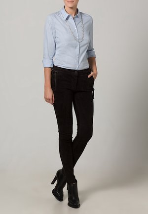 Femme portant une chemise à boutons bleu clair, un pantalon slim noir avec poches zippées, des bottines à talons noirs et un collier chaîne argenté.