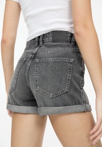Grijze denimshorts met opgerolde zomen, voorzien van achterzakken en een zwart leren label aan de tailleband. Ongewerkte randen en subtiele textuur.