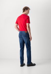 Rdeča prilegajoča majica in modri traper hlače z teksturirano površino. Hlače imajo rjav usnjen znak in dve zadnji žep. Črne čevlje.