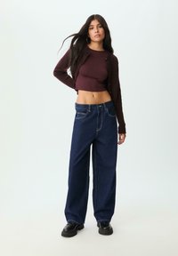 Cardigan lungo a maniche corte color borgogna indossato sopra un top aderente, abbinato a jeans in denim blu scuro larghi con cuciture a contrasto e taglio dritto.
