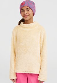 Beiger Fleece-Pullover mit hohem Kragen und langen Ärmeln, ausgestattet mit einer weichen, strukturierten Oberfläche. Kombiniert mit einer pink-blau gestreiften Strickmütze.