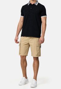 Schwarzes Poloshirt mit gestreiftem Kragen, beigen Cargo-Shorts und weißen Sneakern. Das Shirt verfügt über eine Drei-Knopf-Leiste und ein Logodetail.