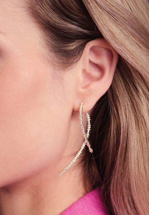 Orecchini lunghi in tono oro con un design intrecciato, decorati con piccole pietre di strass trasparenti. Texture liscia con un'estetica delicata e moderna.