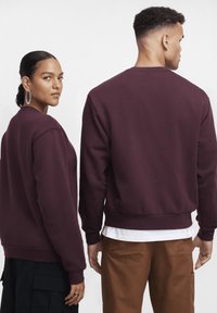 Sweatshirt bordô feita de tecido macio, com gola redonda e punhos canelados. Exibida em duas modelos de costas, combinada com roupas escuras e claras.