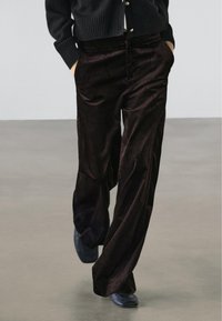 Person iført mørkebrune viddebenet kordfløjlsbukser, sorte sko og en sort, knappet sweater med hænderne i lommerne.